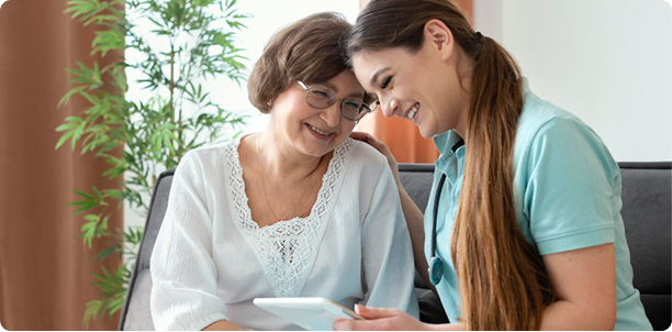 A panoramic medium shot of an older woman with glasses and a white lace-trimmed top sitting on a sofa next to a young female nurse or caregiver wearing a stethoscope. Both are smiling warmly and leaning close together as they look down at a digital tablet held by the caregiver.