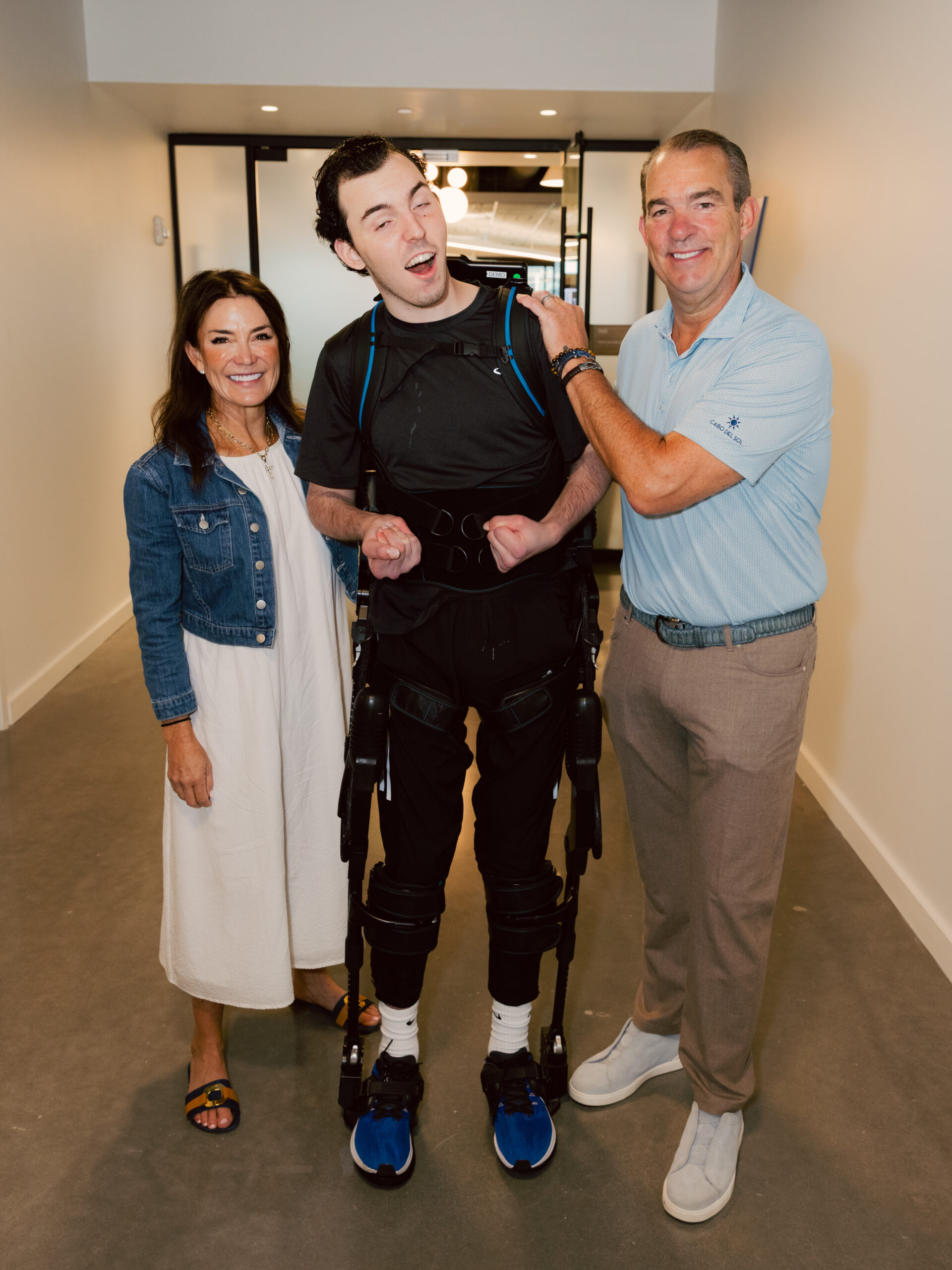 Devon Corbett stands in a hallway supported by a robotic exoskeleton, smiling alongside his parents who stand on either side of him in support.
