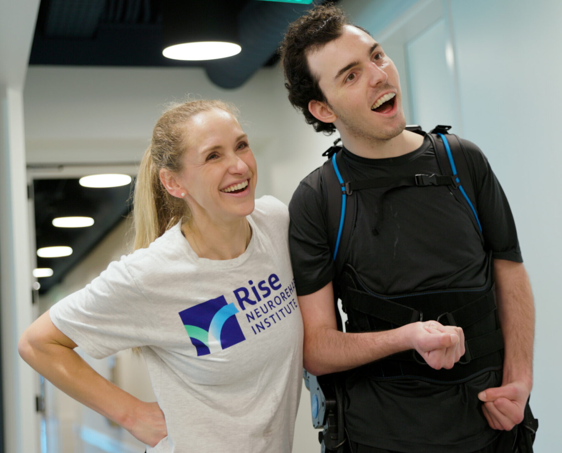 A medium close-up of Devon Corbett standing in a hallway while wearing a black robotic exoskeleton, looking forward with an excited expression. Beside him, his therapist Caitlyn, wearing a 'Rise Neurorehab Institute' t-shirt, stands with her hand on her hip, smiling and laughing along with him.